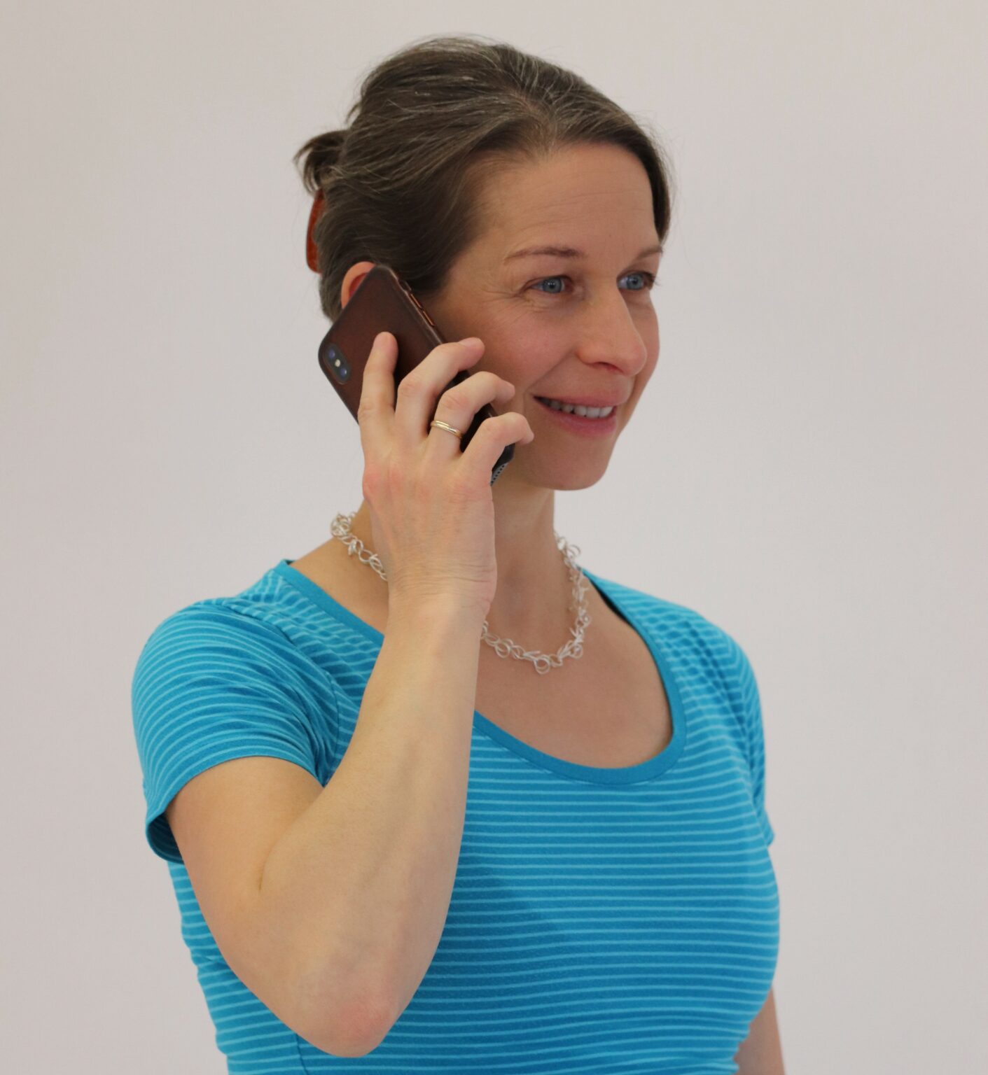 Eine Frau mit zur&uuml;ckgebundenem Haar tr&auml;gt ein blau gestreiftes Oberteil und eine silberne Halskette. Sie steht entspannt und h&auml;lt ein Smartphone an ihr Ohr, w&auml;hrend sie telefoniert. Ihre Haltung wirkt aufrecht und ausgeglichen, was auf ein Bewusstsein f&uuml;r eine ergonomische K&ouml;rperhaltung hinweist. Der Hintergrund ist schlicht und lenkt den Fokus auf die nat&uuml;rliche und entspannte Szene.