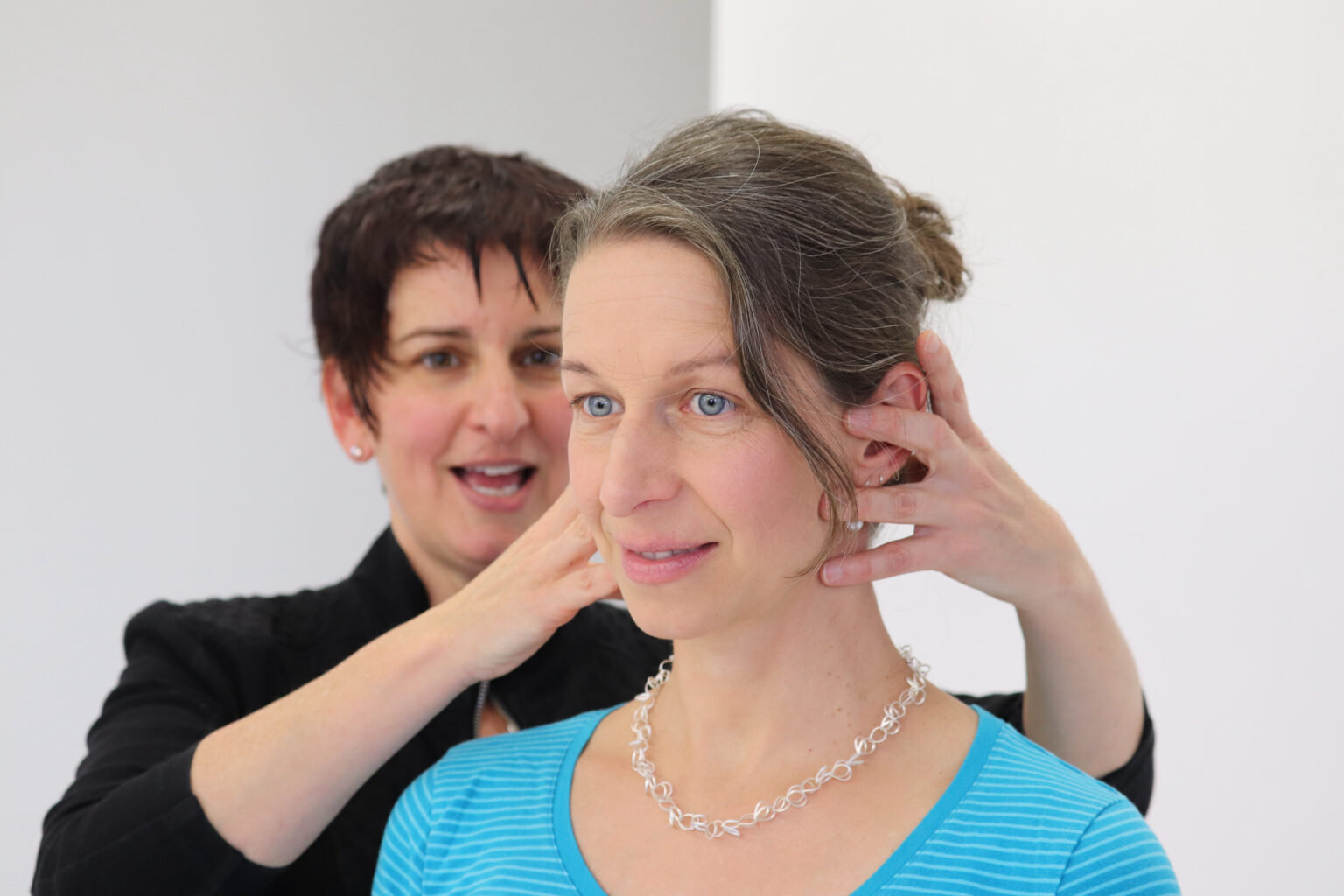 Eine Frau mit kurzen dunklen Haaren und schwarzer Kleidung steht hinter einer anderen Frau mit zur&uuml;ckgebundenem, graumeliertem Haar. Die zweite Frau tr&auml;gt ein blau gestreiftes Oberteil und eine silberne Halskette. Die Frau in Schwarz demonstriert eine Technik der Alexandertechnik, indem sie ihre H&auml;nde sanft an den Kopf und Hals der anderen Frau legt. Der Fokus liegt auf der achtsamen Ber&uuml;hrung und der Verbesserung von Haltung und Bewegung. Der Hintergrund ist schlicht und wei&szlig;, wodurch die Interaktion hervorgehoben wird.
